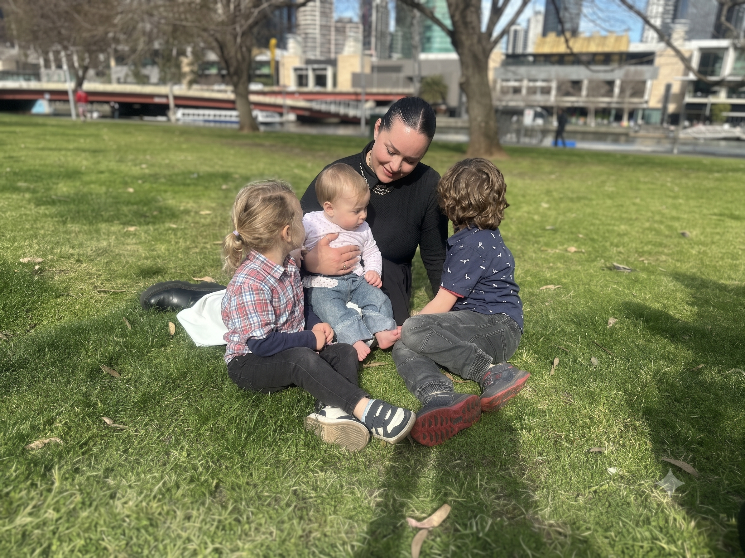 A woman sitting on a grassy park lawn with three children outdoors. The background shows trees, a bridge, and city buildings in soft focus.